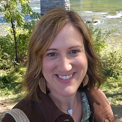 Outdoor headshot of Alyssa Mychaskiw wearing a brown shirt, with trees, a river, and a stone bridge in the background.
